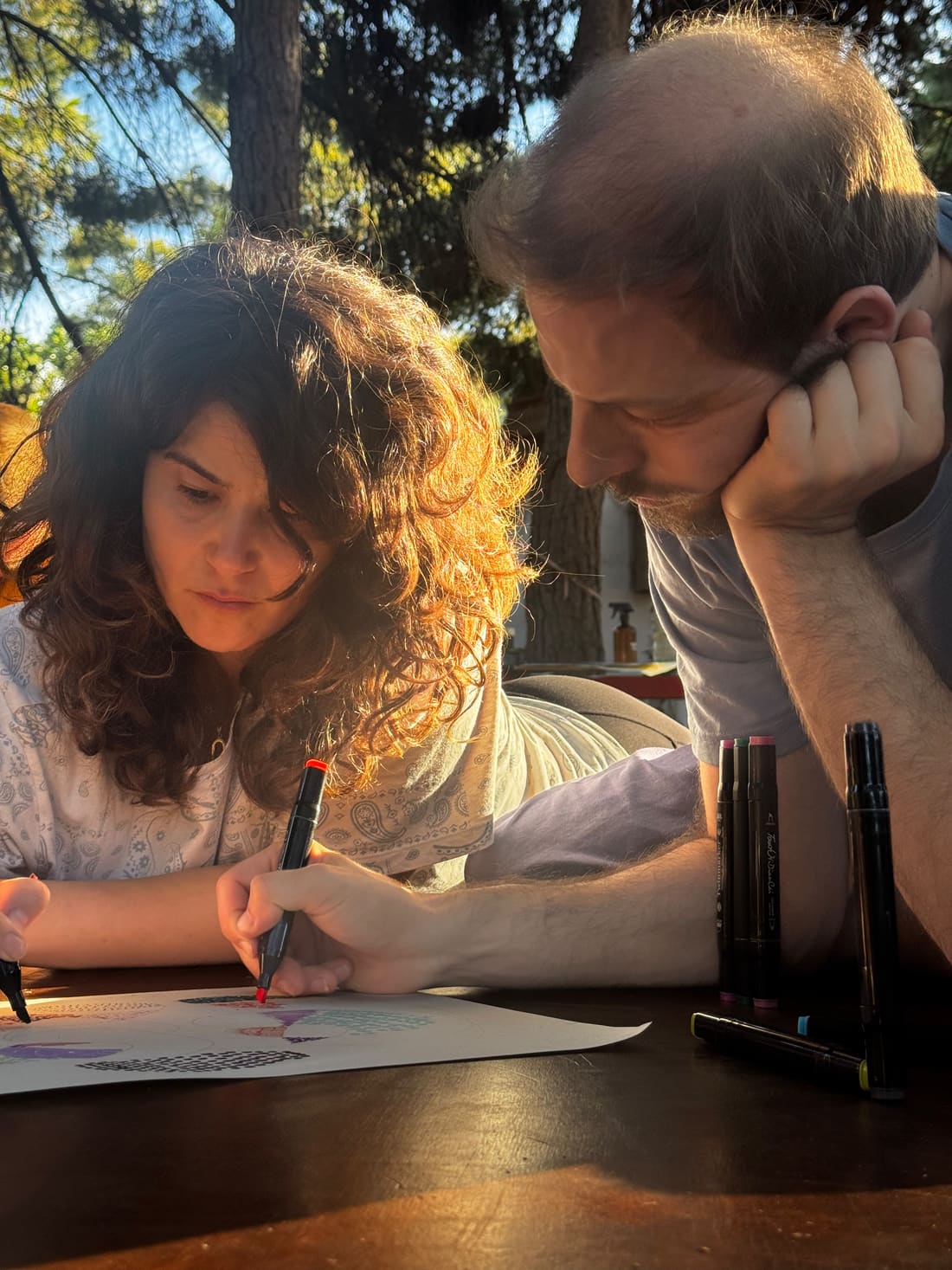 קרין וטל רכונים יחד על דף ציור בחוץ, קרין מציירת בטוש וטל מסתכל, באור שמש של סוף היום